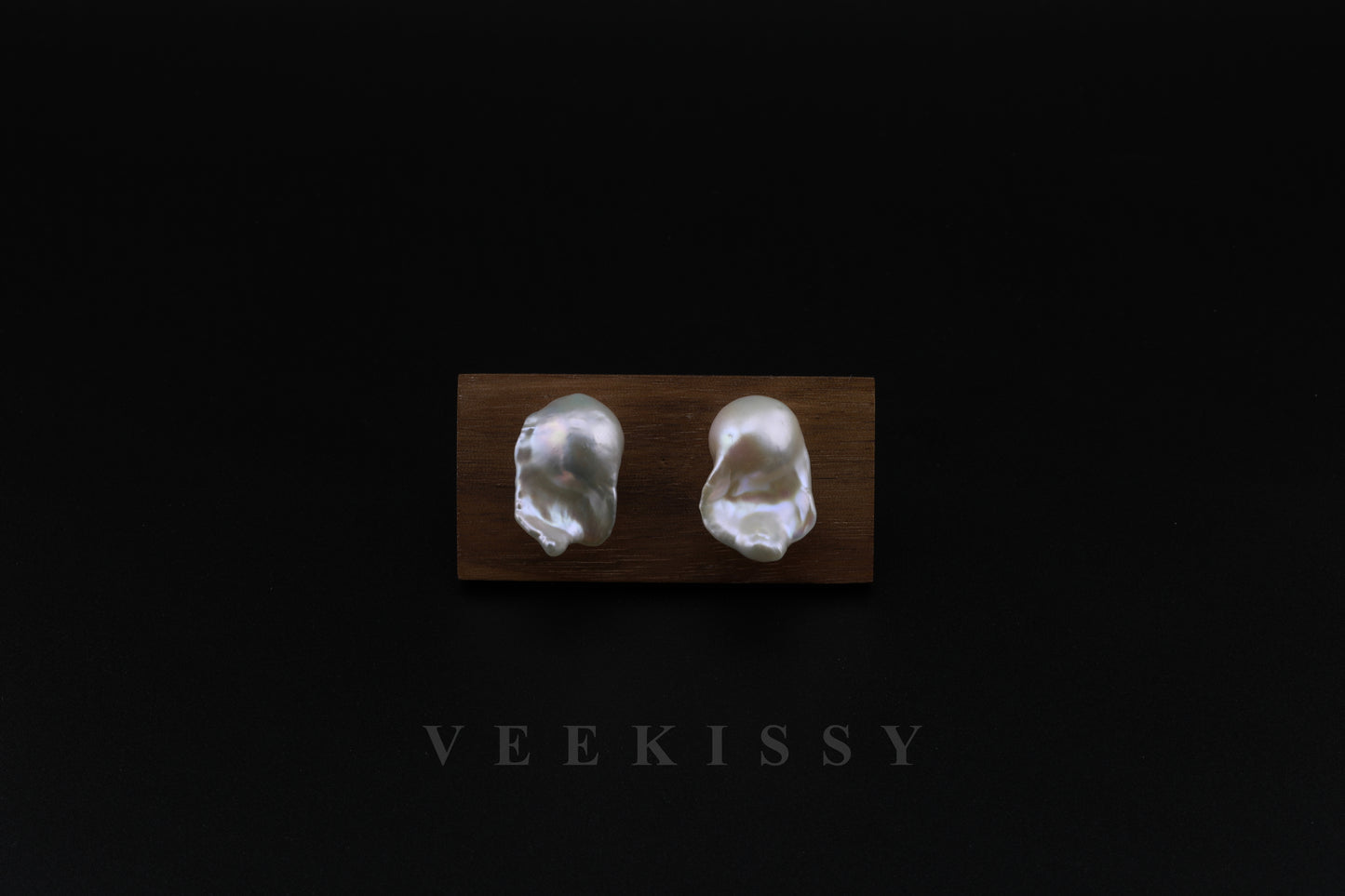 A pair of handcrafted baroque pearl stud earrings displayed on a wooden stand against a black background, featuring natural freshwater pearls with a unique organic shape and sterling silver studs.