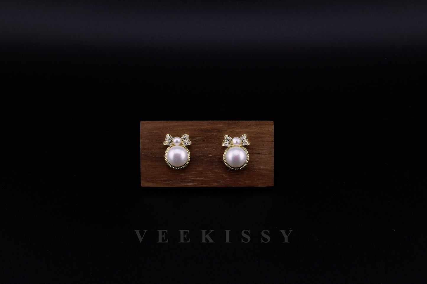 A pair of elegant freshwater pearl stud earrings with a delicate gold-plated sterling silver bow design, set against a dark background on a wooden display. Handcrafted by VEEKISSY in Sydney