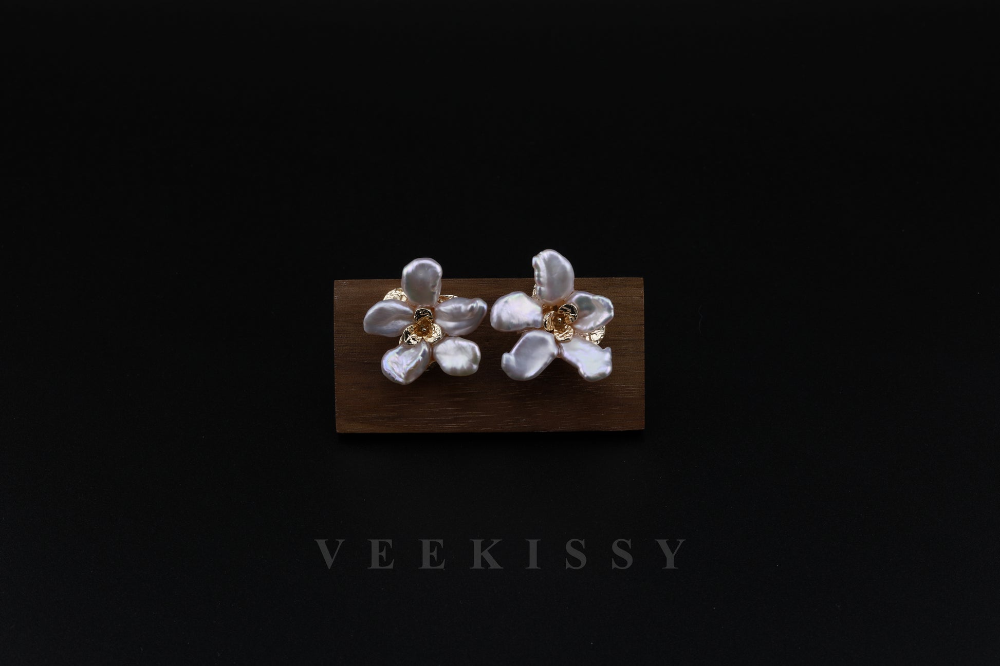 A pair of handcrafted floral earrings featuring baroque pearl petals set in gold-plated sterling silver, displayed against a black background with a wooden stand.