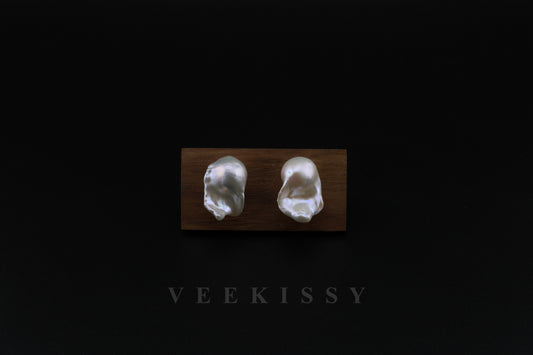 A pair of handcrafted baroque pearl stud earrings displayed on a wooden stand against a black background, featuring natural freshwater pearls with a unique organic shape and sterling silver studs.