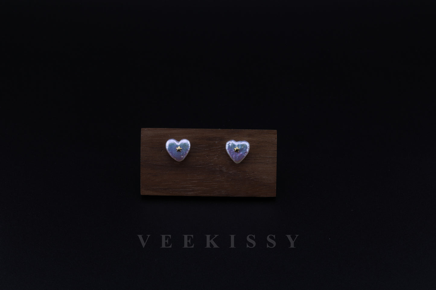 A pair of heart-shaped baroque pearl stud earrings with a delicate 18K gold star accent, displayed on a wooden stand against a black background.