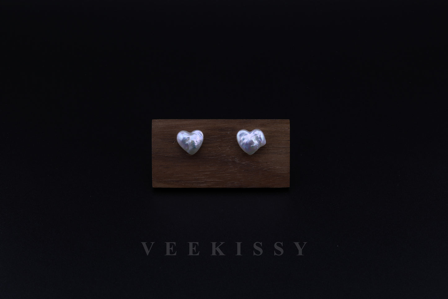 A pair of handcrafted heart-shaped baroque pearl earrings displayed on a wooden stand against a black background, featuring genuine freshwater pearls set on sterling silver studs.