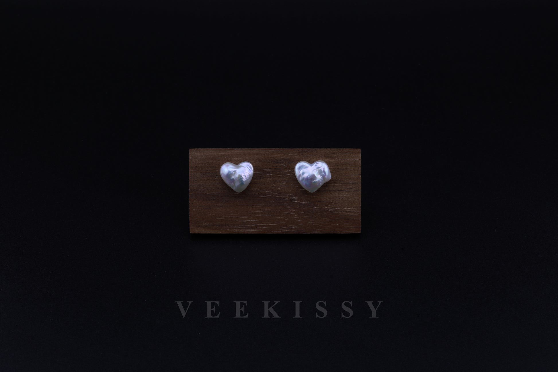 A pair of handcrafted heart-shaped baroque pearl earrings displayed on a wooden stand against a black background, featuring genuine freshwater pearls set on sterling silver studs.