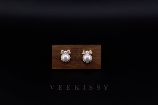 A pair of elegant freshwater pearl stud earrings with a delicate gold-plated sterling silver bow design, set against a dark background on a wooden display. Handcrafted by VEEKISSY in Sydney