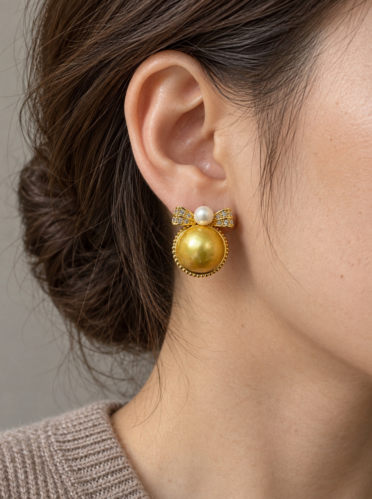 Golden bow pearl stud earring worn on the ear with taupe knitwear